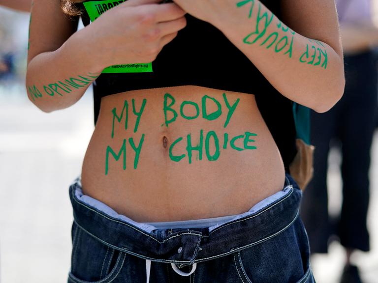 Eine Frau mit bauchfreiem Top nimmt an einer Demonstration teil. Auf ihrem Bauch sind mit grüner Farbe die Wörter "my body my choice" zu lesen.