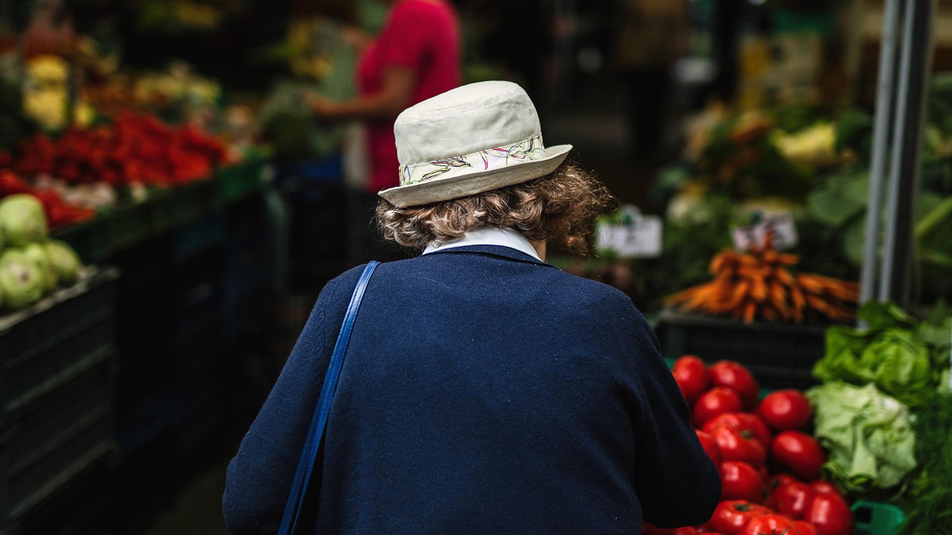 Eine ältere Frau mit Hut und dem Rücken zur Kamera geht über einen Gemüsemarkt. 
