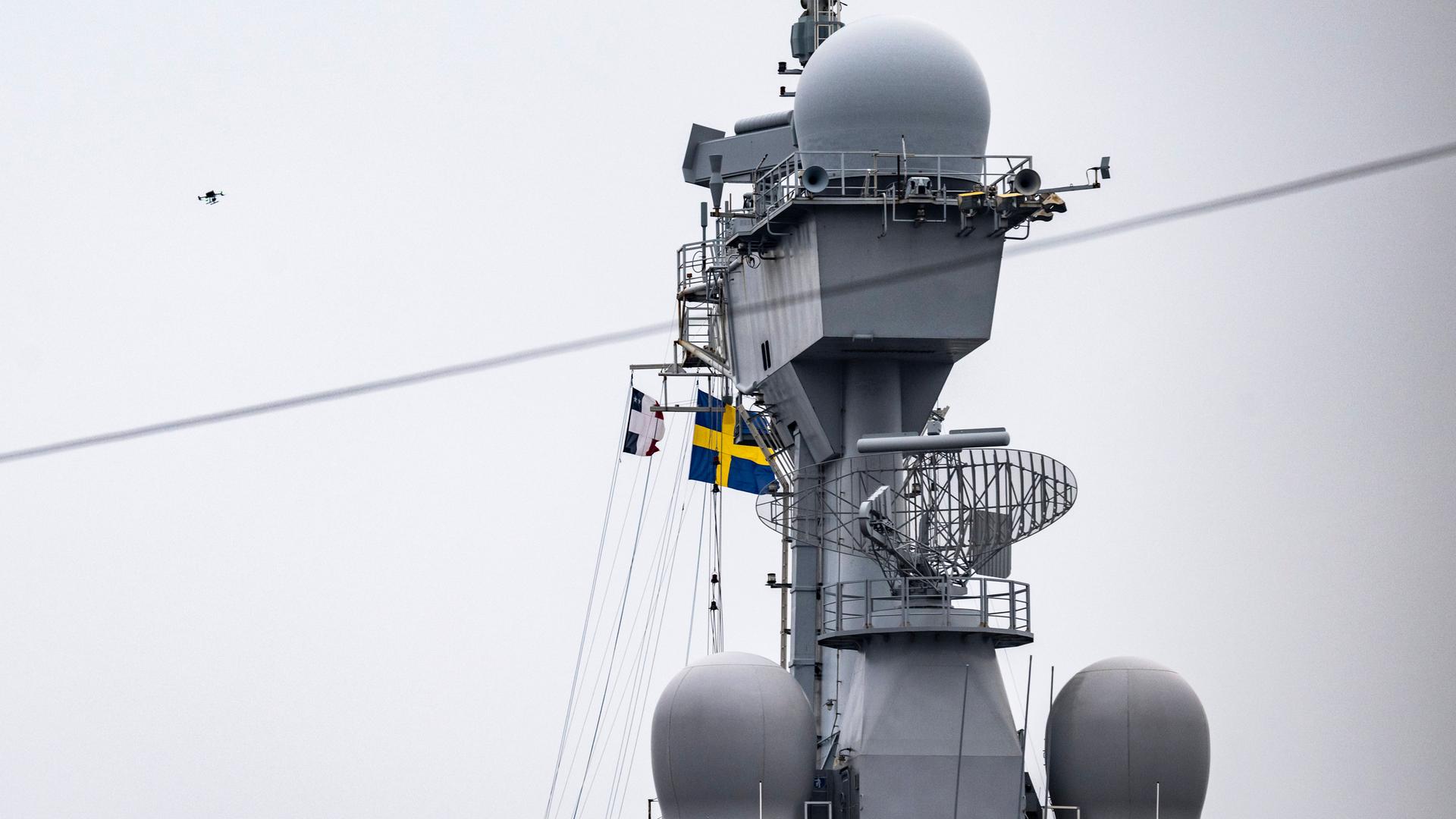 Ein graues Schiff in einem Hafen, im Hintergrund ist unscharf eine Drohne zu erkennen und die schwedische Flagge in blau und gelb. Ein graues Schiff in einem Hafen, im Hintergrund ist unscharf eine Drohne zu erkennen und die schwedische Flagge in blau und gelb.