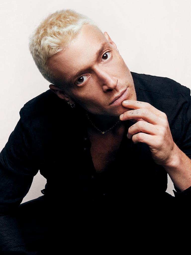 Portrait eines jungen Mannes, mit kurzen blondierten Haaren, der die Finger seiner linken Hand am Kinn hält und den Betrachter anblickt.