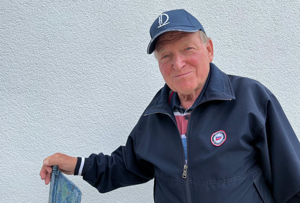 Ein alter Mann blauer Mütze und blauer Jacke steht vor einer Hauswand.