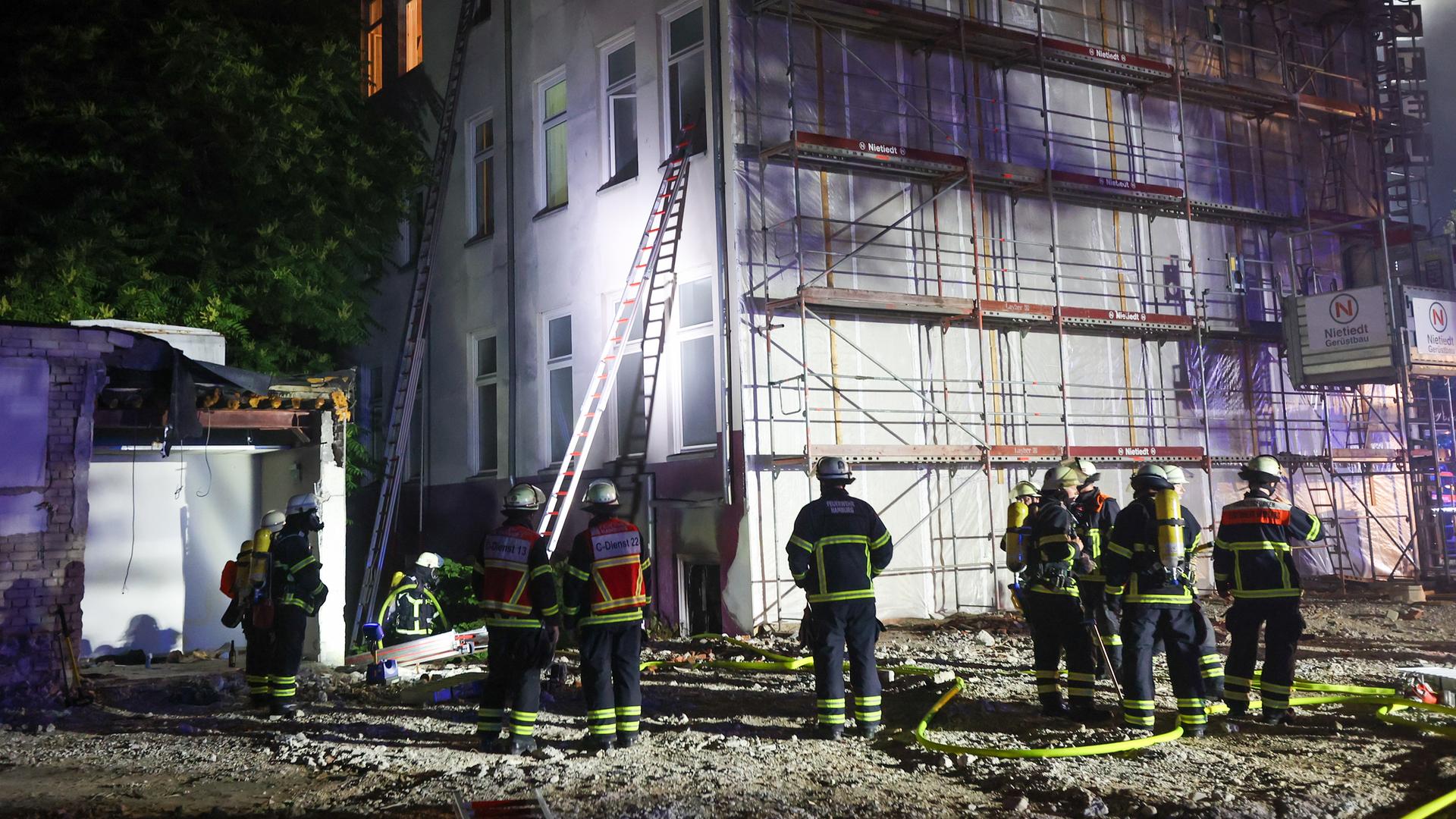 Feuerwehrleute stehen in der Nacht vor einem brennenden Gebäude. Zwei Leitern führen zu Fenstern. Feuerwehrleute stehen in der Nacht vor einem brennenden Gebäude. Zwei Leitern führen zu Fenstern.