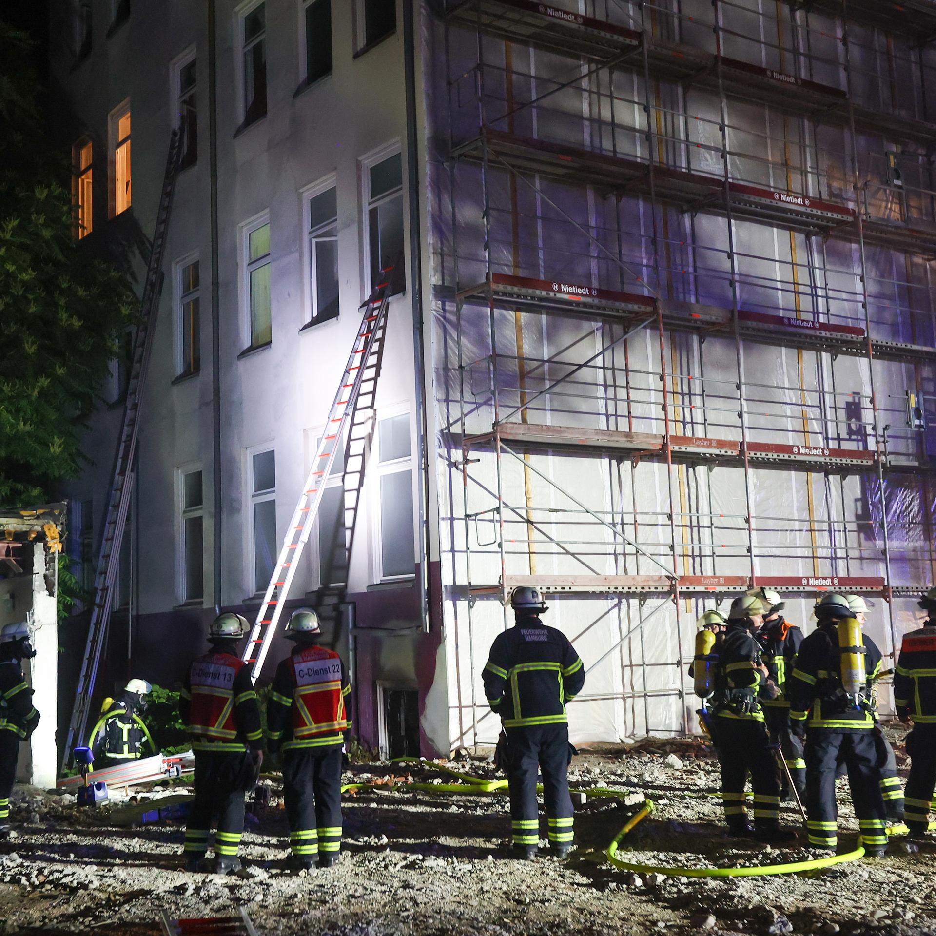 Feuerwehrleute stehen in der Nacht vor einem brennenden Gebäude. Zwei Leitern führen zu Fenstern.