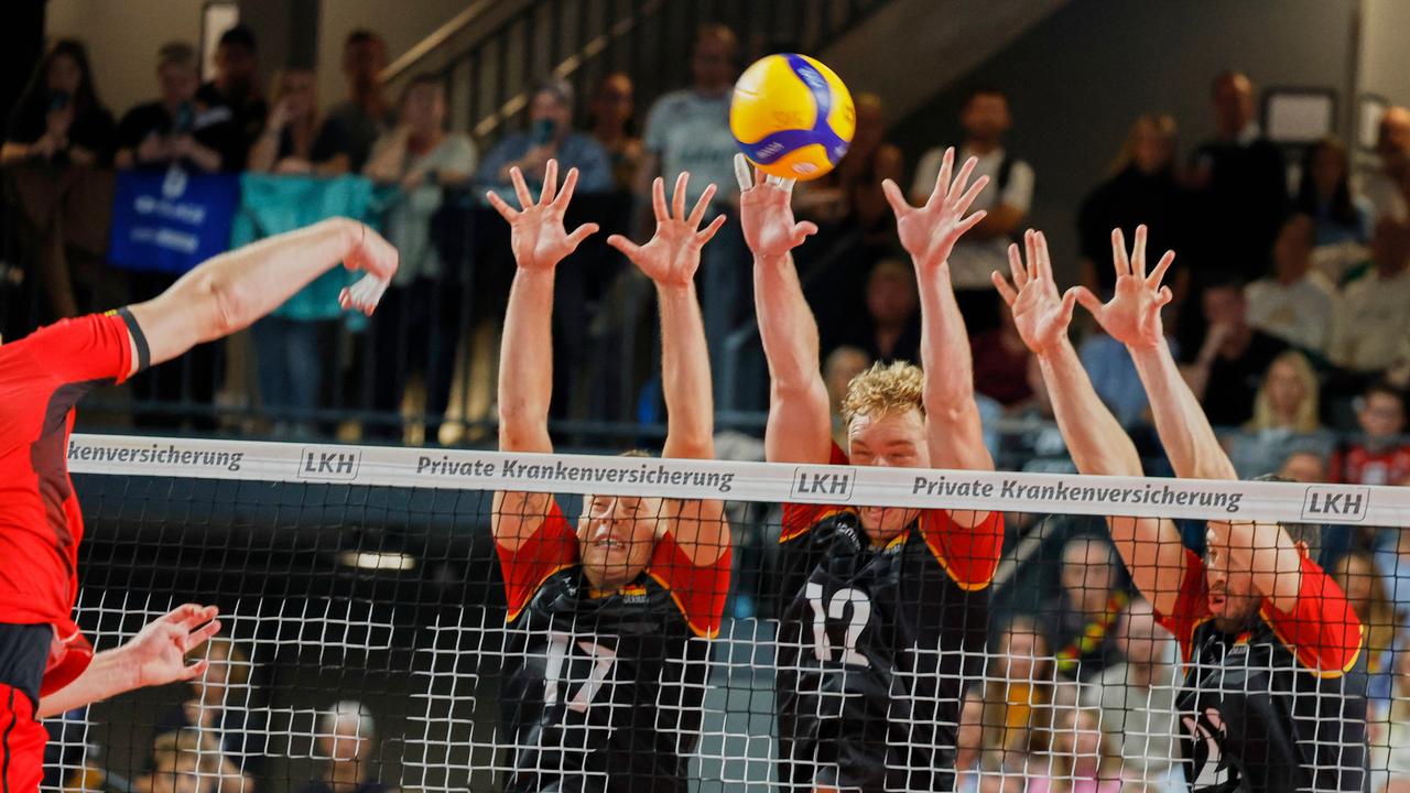 Drei Männer im Trikot der deutschen Volleyball-Nationalmannschaft reißen am Netz die Arme hoch, um einen Ball abzuwehren. 