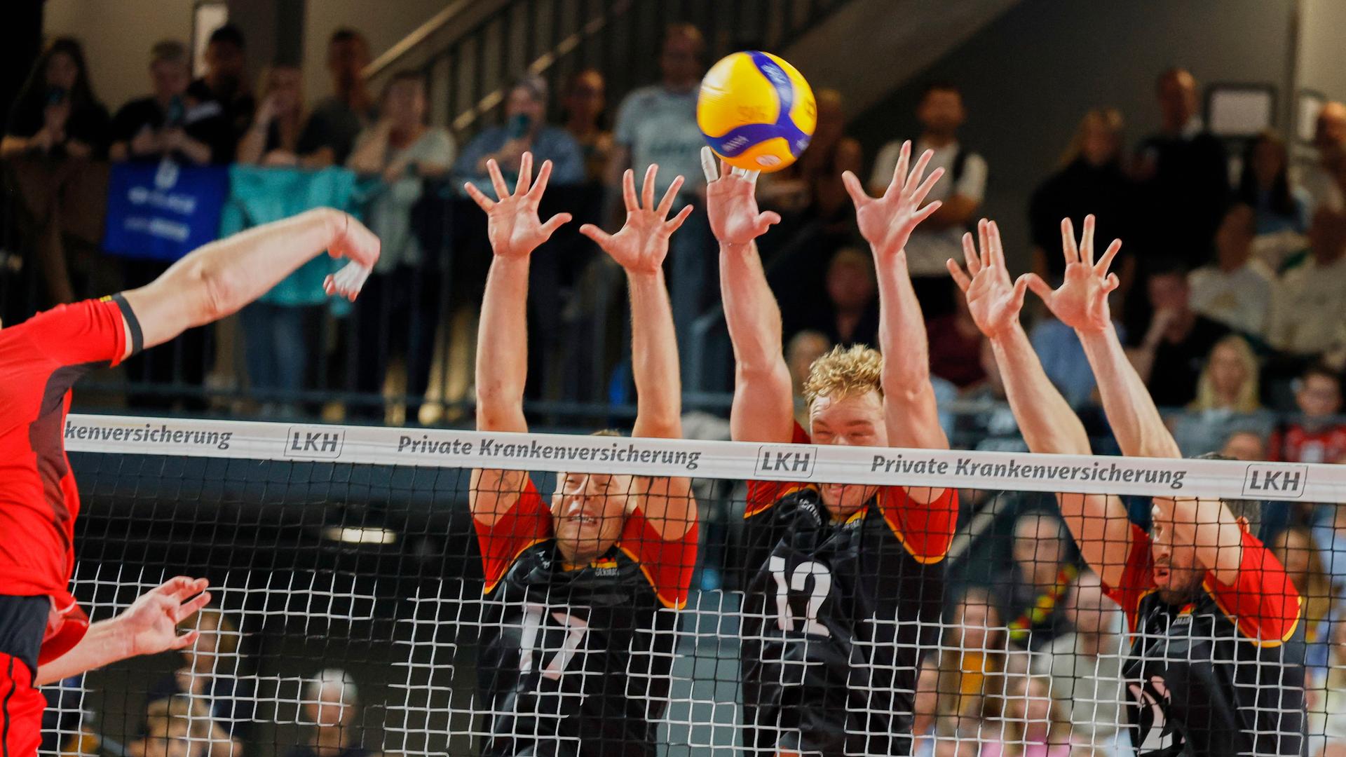 Drei Männer im Trikot der deutschen Volleyball-Nationalmannschaft reißen am Netz die Arme hoch, um einen Ball abzuwehren. 