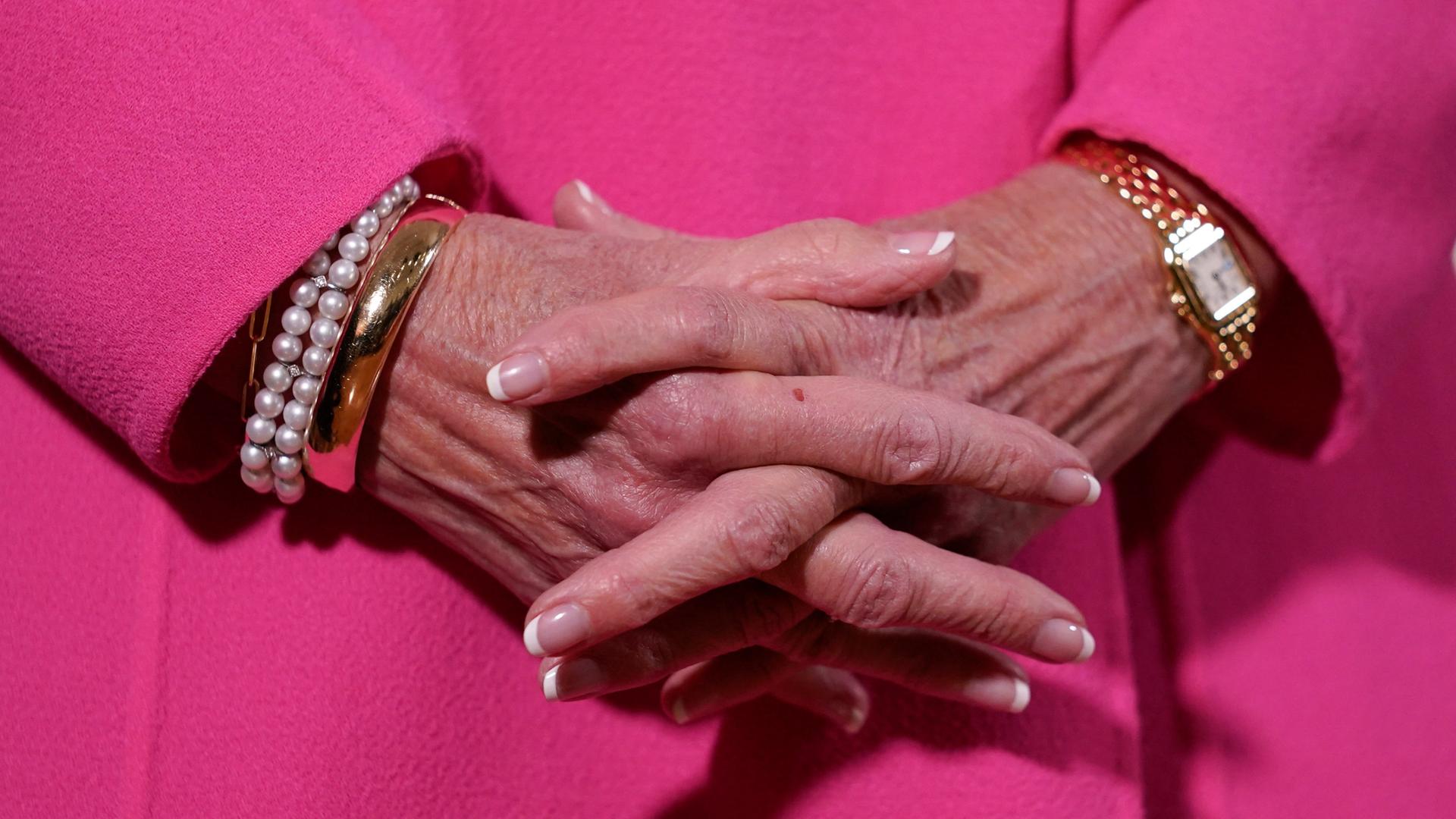 Die Hände von US-First Lady Jill Biden sind vor einem magentafarbenen Mantel zusammengefaltet. An dem einen Arm ist Schmuck, am anderen eine Uhr. 