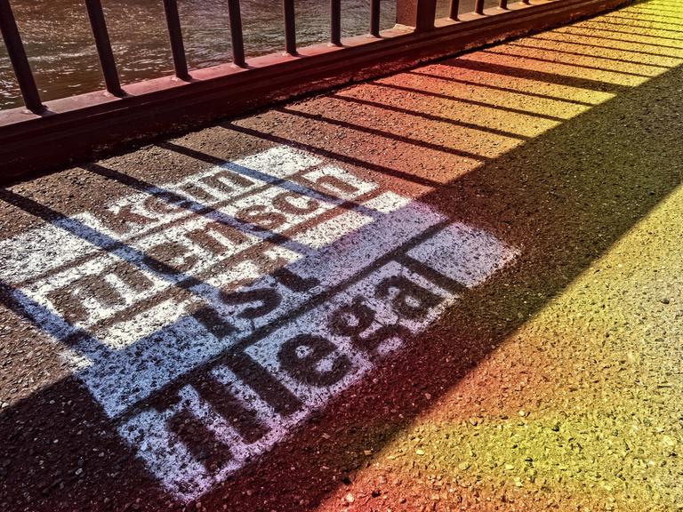 Auf den Asphalt vor einem Zaun ist mit weißer Farbe der Schriftzug gesprüht: "Kein Mensch ist illegal".
