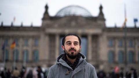Der Grünen-Politiker Tareq Alaows steht vor dem Bundestag in Berlin und schaut seitwärts an der Kamera vorbei. 