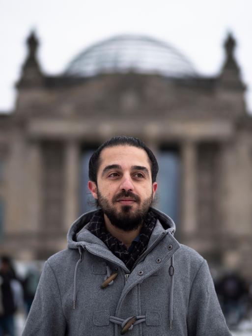 Der Grünen-Politiker Tareq Alaows steht vor dem Bundestag in Berlin und schaut seitwärts an der Kamera vorbei. 