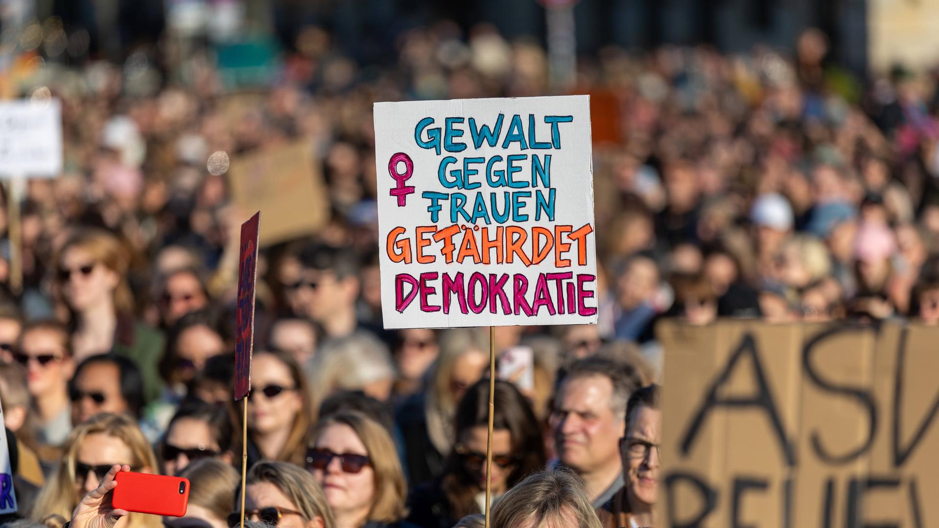 Zahlreiche Menschen protestieren auf der Straße gegen sexualisierte digitale Gewalt. Eine Person hält ein Schild hoch mit der Aufschrift "Gewalt gegen Frauen gefährdet Demokratie" Zahlreiche Menschen protestieren auf der Straße gegen sexualisierte digitale Gewalt. Eine Person hält ein Schild hoch mit der Aufschrift "Gewalt gegen Frauen gefährdet Demokratie"