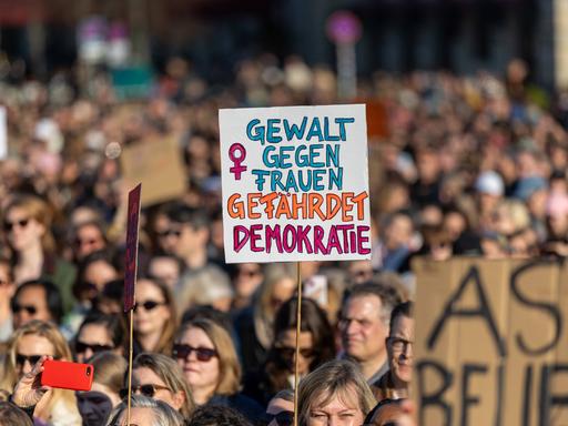 Zahlreiche Menschen protestieren auf der Straße gegen sexualisierte digitale Gewalt. Eine Person hält ein Schild hoch mit der Aufschrift "Gewalt gegen Frauen gefährdet Demokratie" Zahlreiche Menschen protestieren auf der Straße gegen sexualisierte digitale Gewalt. Eine Person hält ein Schild hoch mit der Aufschrift "Gewalt gegen Frauen gefährdet Demokratie"