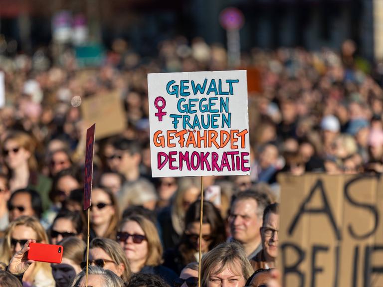 Zahlreiche Menschen protestieren auf der Straße gegen sexualisierte digitale Gewalt. Eine Person hält ein Schild hoch mit der Aufschrift "Gewalt gegen Frauen gefährdet Demokratie"