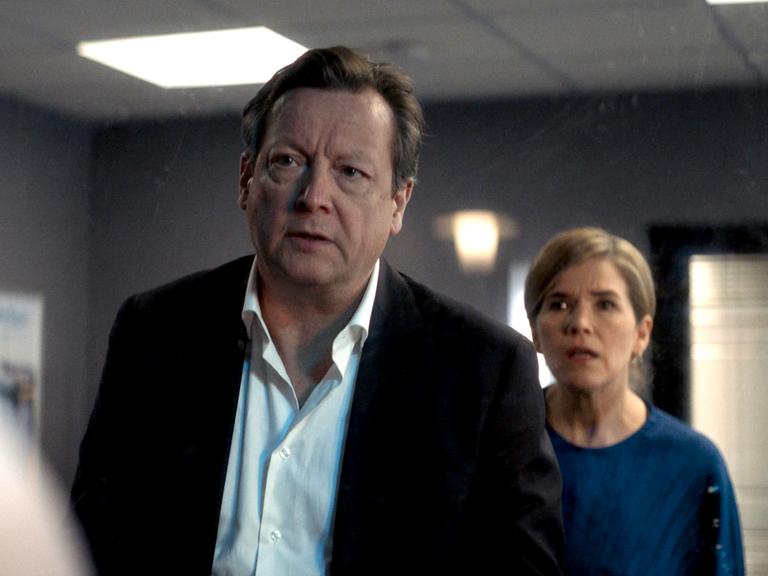 Das Szenenfoto aus dem Film "Kurzschluss" zeigt den Schauspieler Matthias Brandt und die Schauspielerin Anke Angelke, die mit grimmigem Blick hintereinander in einem wenig beleuchteten Raum stehen.