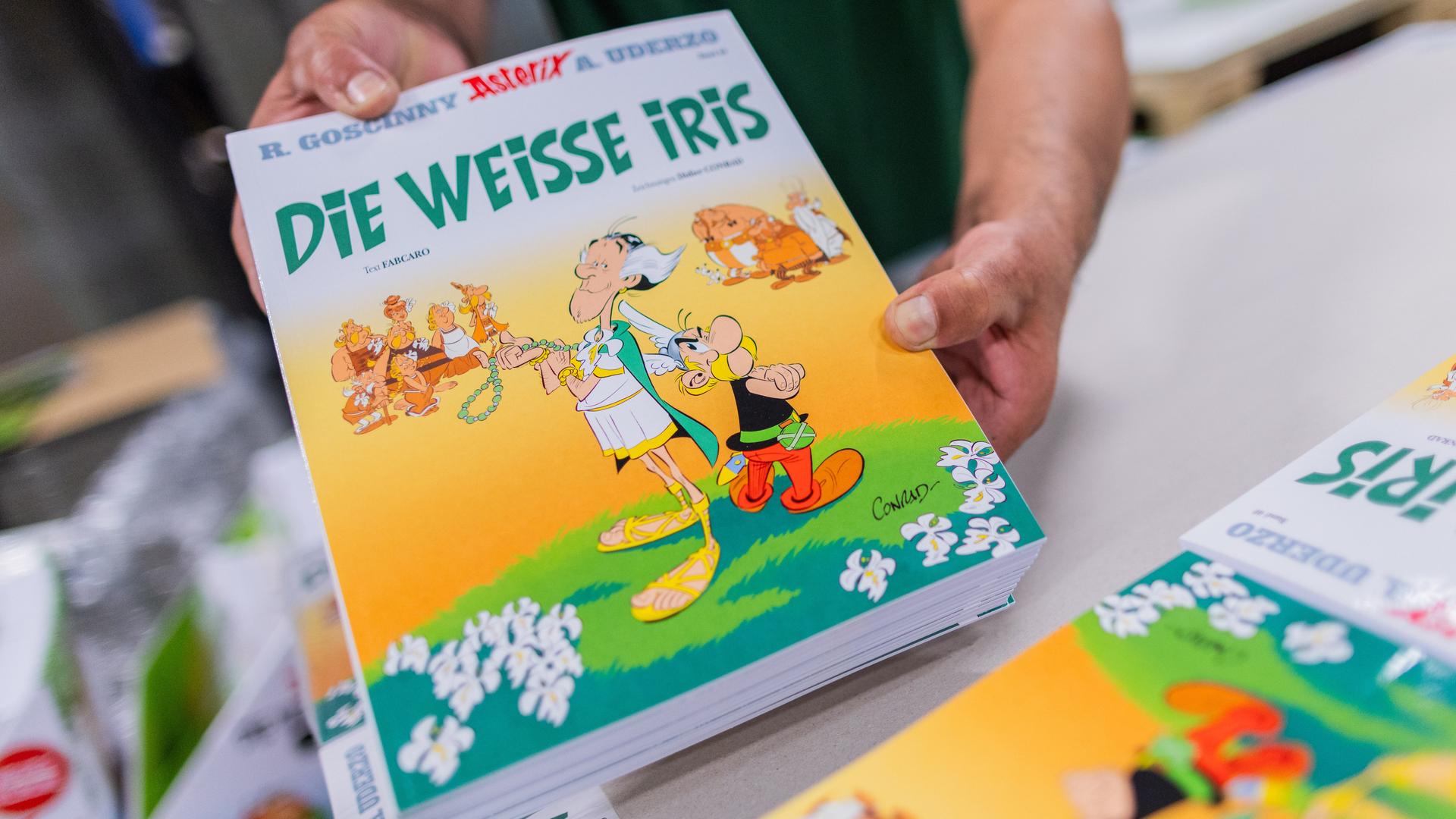 Ein Mann hält mehrere Ausgaben des neuen Asterix-Bandes in Händen und zeigt sie in die Kamera. Weitere Ausgaben liegen auf einem Tisch.