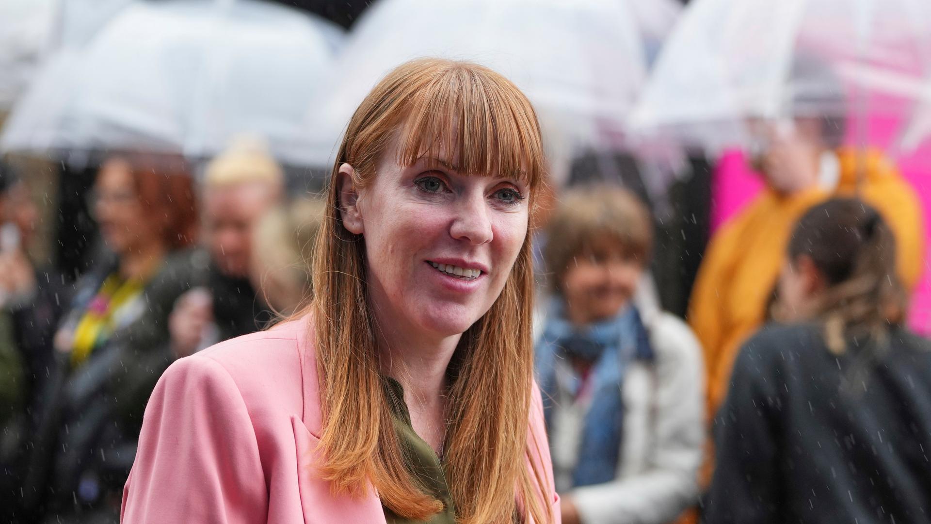 Angela Rayner lächelt in die Kamera. Hinter ihr sieht man verschwommen Menschen, die Regenschirme halten. Angela Rayner lächelt in die Kamera. Hinter ihr sieht man verschwommen Menschen, die Regenschirme halten.