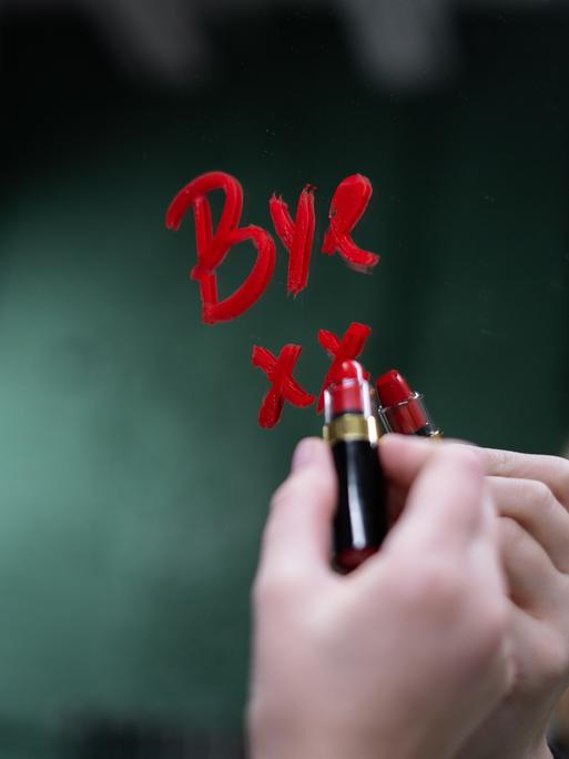 Eine Hand schreibt mit Lippenstift "Bye" auf einen Spiegel