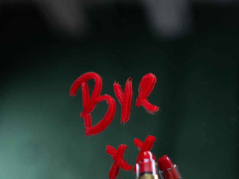 Eine Hand schreibt mit Lippenstift "Bye" auf einen Spiegel