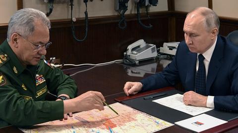 Russlands Verteidigungsminister Schoigu sitzt an einem Tisch und zeigt mit einem Stift auf eine Karte. Rechts neben ihm sitzt Präsident Putin.