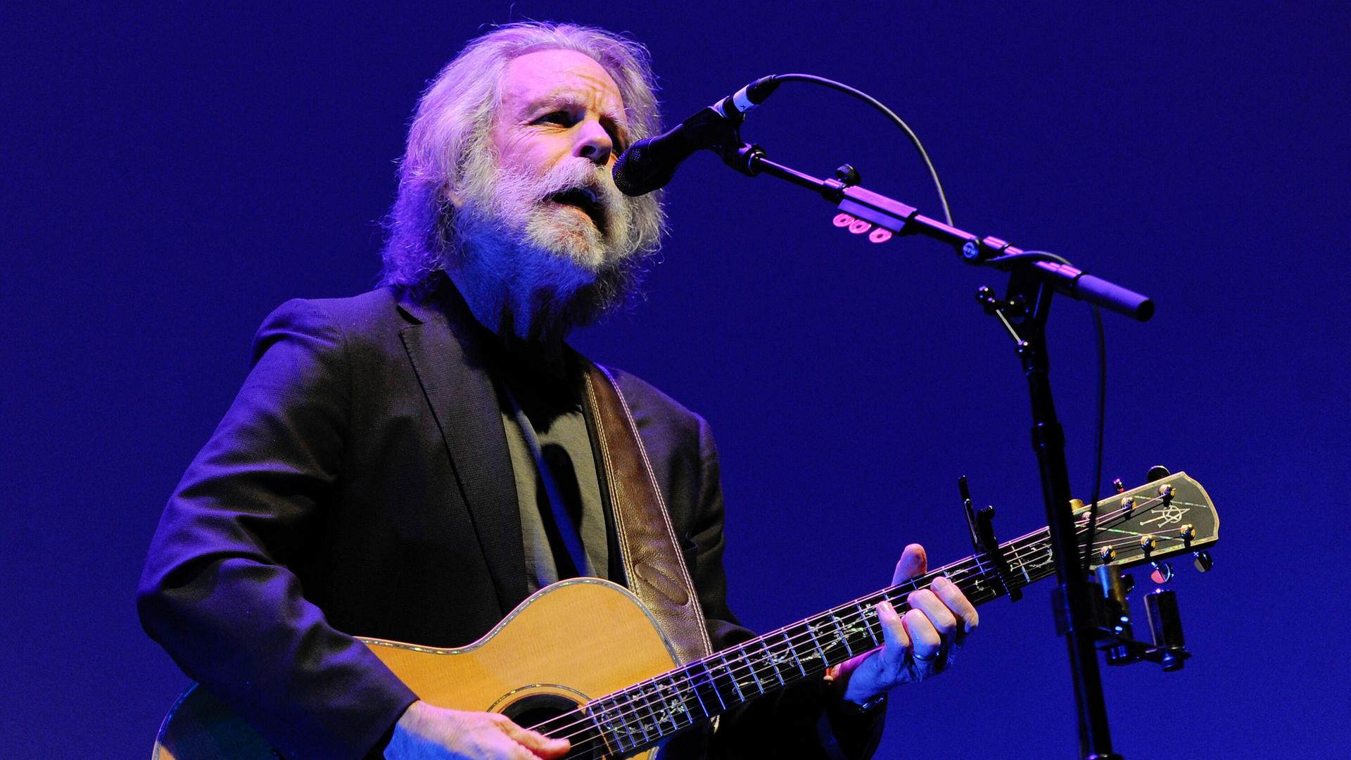Weir steht auf einer Bühne am Mikrofon, singt und spielt eine Akustikgitarre. Er trägt ein schwarzes Jackett und hat graue Haare sowie einen grauen Rauschebart. Der Hintergrund ist blau.