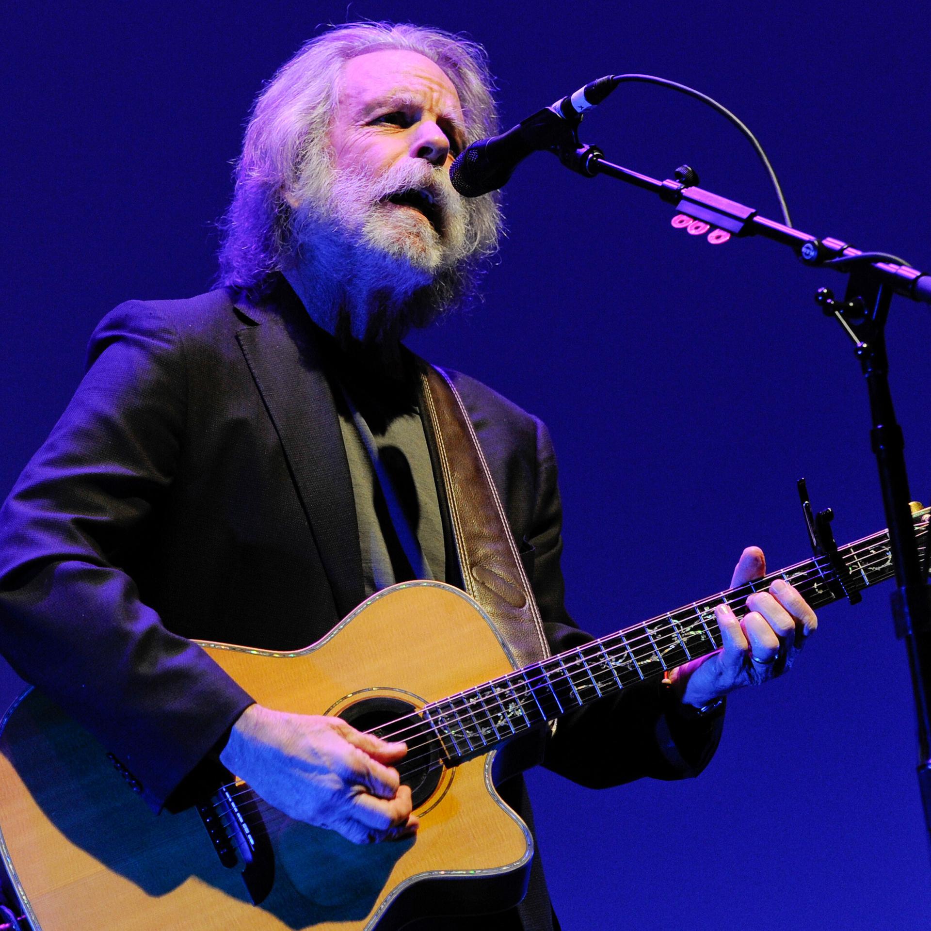 Weir steht auf einer Bühne am Mikrofon, singt und spielt eine Akustikgitarre. Er trägt ein schwarzes Jackett und hat graue Haare sowie einen grauen Rauschebart. Der Hintergrund ist blau.