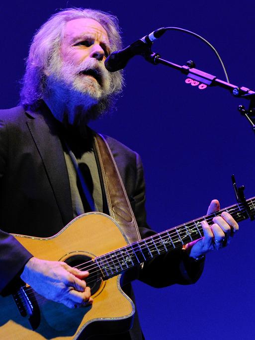 Weir steht auf einer Bühne am Mikrofon, singt und spielt eine Akustikgitarre. Er trägt ein schwarzes Jackett und hat graue Haare sowie einen grauen Rauschebart. Der Hintergrund ist blau.