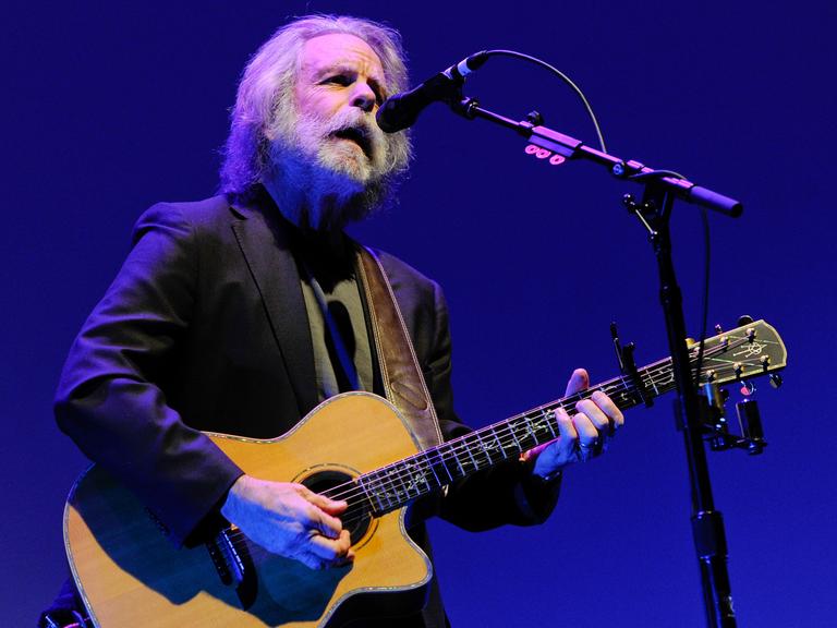 Weir steht auf einer Bühne am Mikrofon, singt und spielt eine Akustikgitarre. Er trägt ein schwarzes Jackett und hat graue Haare sowie einen grauen Rauschebart. Der Hintergrund ist blau.