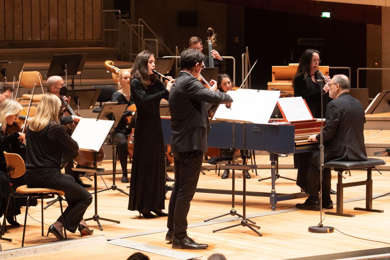 Auf einer Bühne ist ein festlich gekleidetes Orchester zu sehen, auf der rechts ein Mann am Cembalo nach hinten gewendet zu den Musikern spielt. Vorne spielen eine Flötistin und ein Geiger.