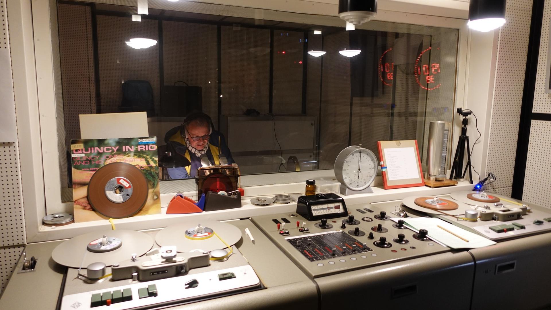 Ein Mann spricht hinter einer Scheibe in ein Mikrofon, davor ist alte Sendetechnik in einem Studio zu sehen