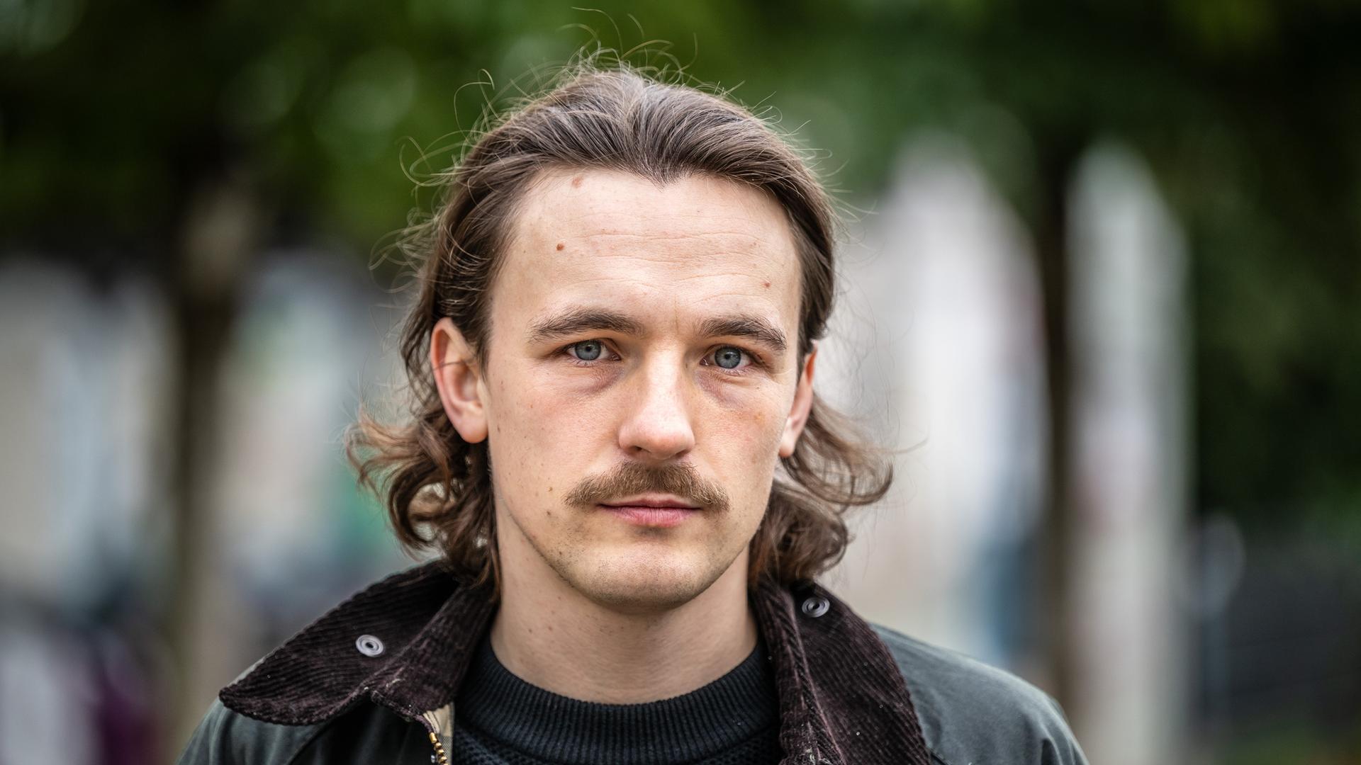 Der Autor Kaleb Erdmann steht auf dem Fürstenplatz in Düsseldorf. Er trägt einen Schnurrbart und eine schwarze Lederjacke.