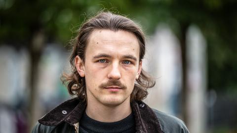 Der Autor Kaleb Erdmann steht auf dem Fürstenplatz in Düsseldorf. Er trägt einen Schnurrbart und eine schwarze Lederjacke. Der Autor Kaleb Erdmann steht auf dem Fürstenplatz in Düsseldorf. Er trägt einen Schnurrbart und eine schwarze Lederjacke.