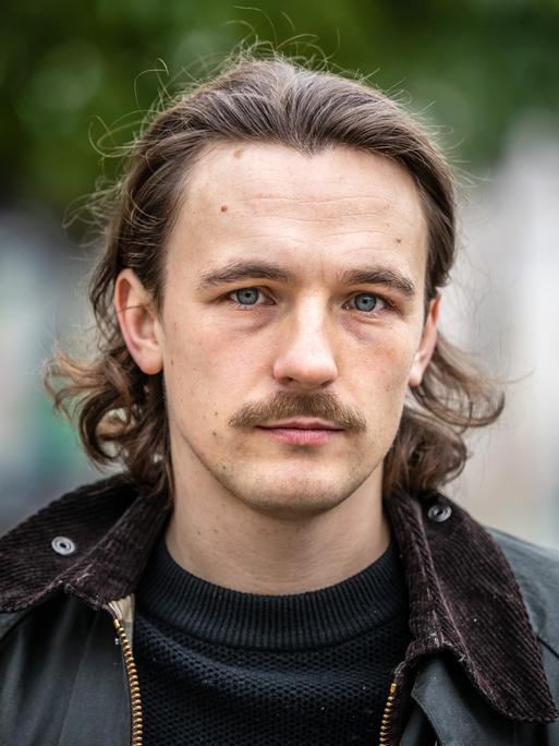 Der Autor Kaleb Erdmann steht auf dem Fürstenplatz in Düsseldorf. Er trägt einen Schnurrbart und eine schwarze Lederjacke.