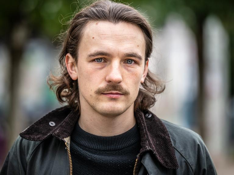 Der Autor Kaleb Erdmann steht auf dem Fürstenplatz in Düsseldorf. Er trägt einen Schnurrbart und eine schwarze Lederjacke.