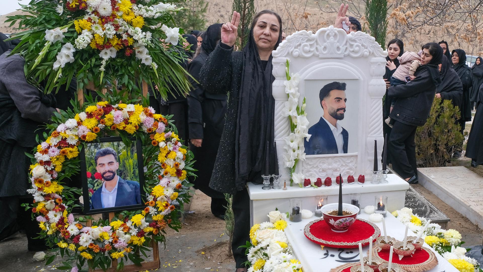 Das Grab eines jungen Mannes ist mit Blumen und Kerzen geschmückt, seine Mutter formt mit beiden Händen das Voctory-Zeichen neben dem Grabstein, der sein Porträtfoto zeigt.