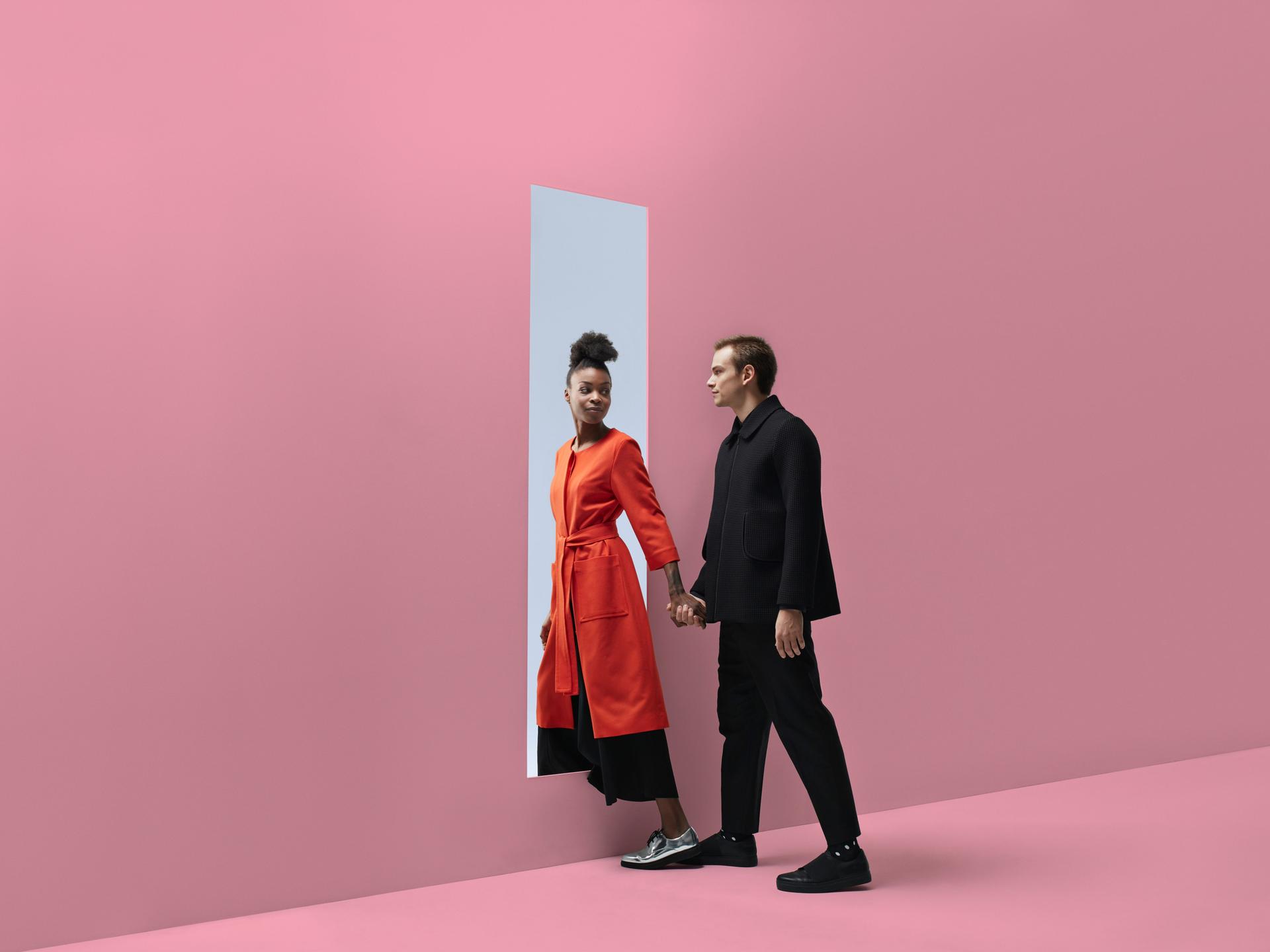 Zwei Personen, die sich an der Hand halten, verlassen einen Raum durch ein rechteckiges Loch in einer pinkfarbenen Wand. Dahinter ist blauer Himmel zu sehen.
