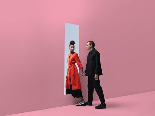 Zwei Personen, die sich an der Hand halten, verlassen einen Raum durch ein rechteckiges Loch in einer pinkfarbenen Wand. Dahinter ist blauer Himmel zu sehen.