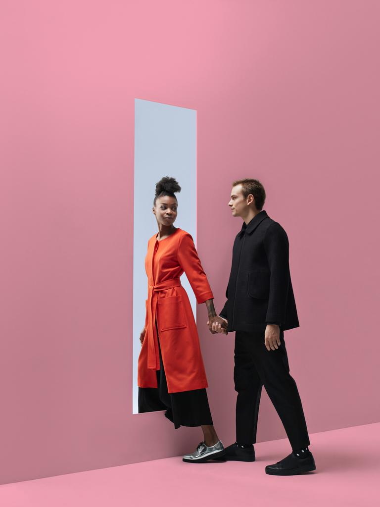Zwei Personen, die sich an der Hand halten, verlassen einen Raum durch ein rechteckiges Loch in einer pinkfarbenen Wand. Dahinter ist blauer Himmel zu sehen.
