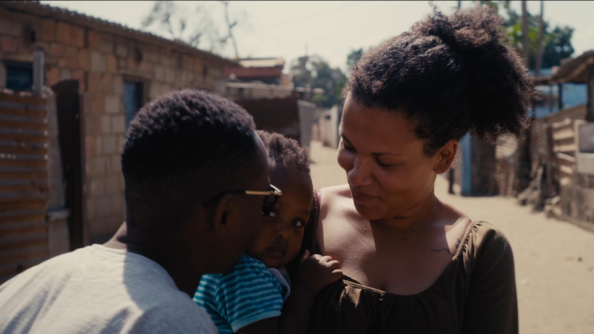 Filmszene aus dem Dokumentarfilm "The Homes We Carry". Zu sehen ist eine junge mosambikanische Frau (vermutlich die Protagonistin des Film) mit Kind auf dem Arm sowie die Rückansicht eines Mannes.