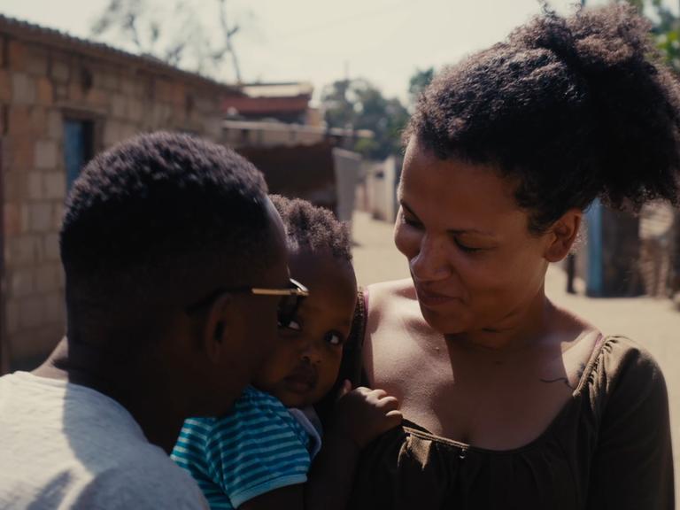 Filmszene aus dem Dokumentarfilm "The Homes We Carry". Zu sehen ist eine junge mosambikanische Frau (vermutlich die Protagonistin des Film) mit Kind auf dem Arm sowie die Rückansicht eines Mannes.