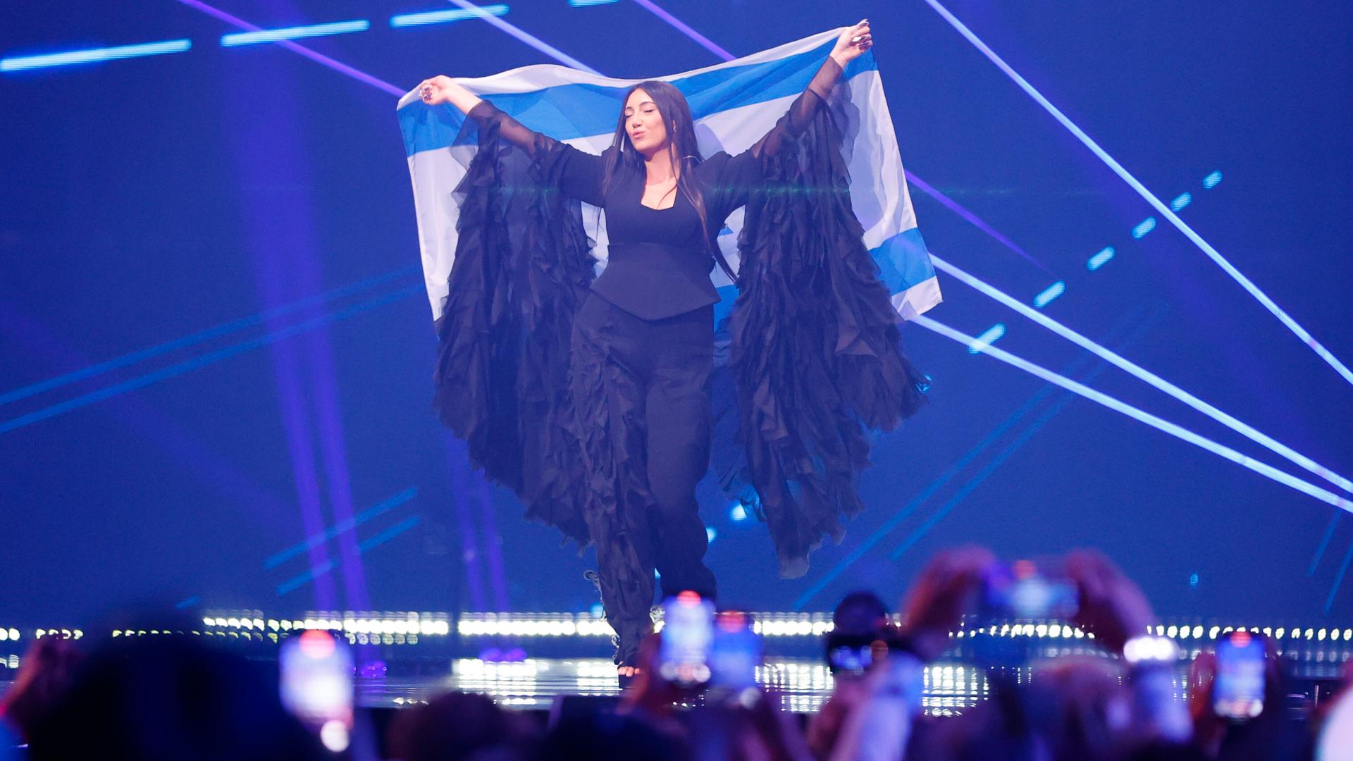 Die israelische Sängerin Yuval Raphael beim ESC. Sie steht auf der Bühne mit blauem Hintergrund und hält eine israelische Fahne hoch. Die israelische Sängerin Yuval Raphael beim ESC. Sie steht auf der Bühne mit blauem Hintergrund und hält eine israelische Fahne hoch.