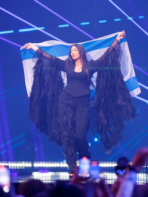 Die israelische Sängerin Yuval Raphael beim ESC. Sie steht auf der Bühne mit blauem Hintergrund und hält eine israelische Fahne hoch. 