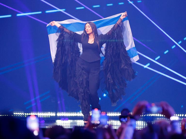 Die israelische Sängerin Yuval Raphael beim ESC. Sie steht auf der Bühne mit blauem Hintergrund und hält eine israelische Fahne hoch. 