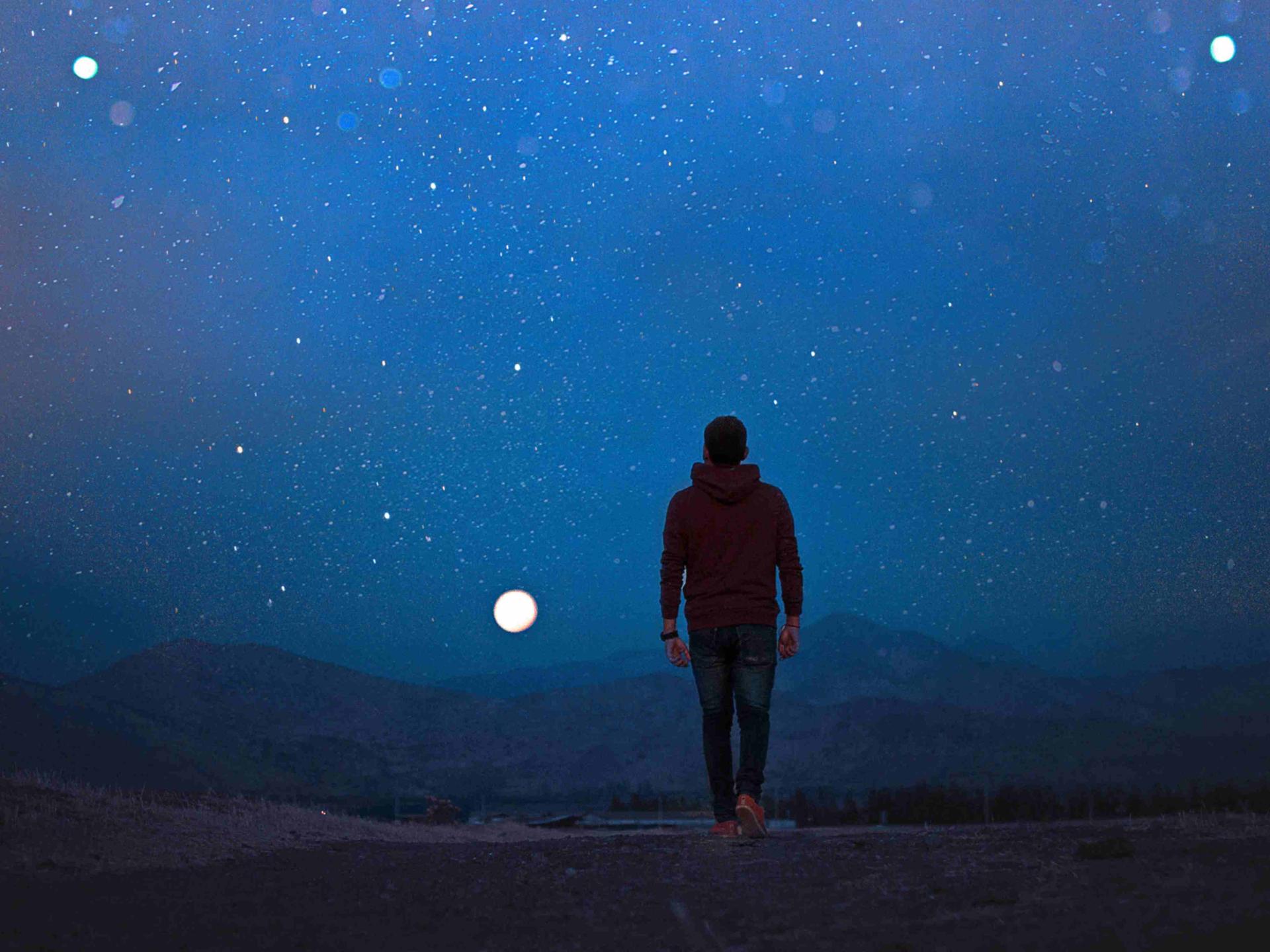 Ein Mann, mit dem Rücken zur Kamera, geht im Dunkeln in einer einsamen Landschaft dem Vollmond entgegen.