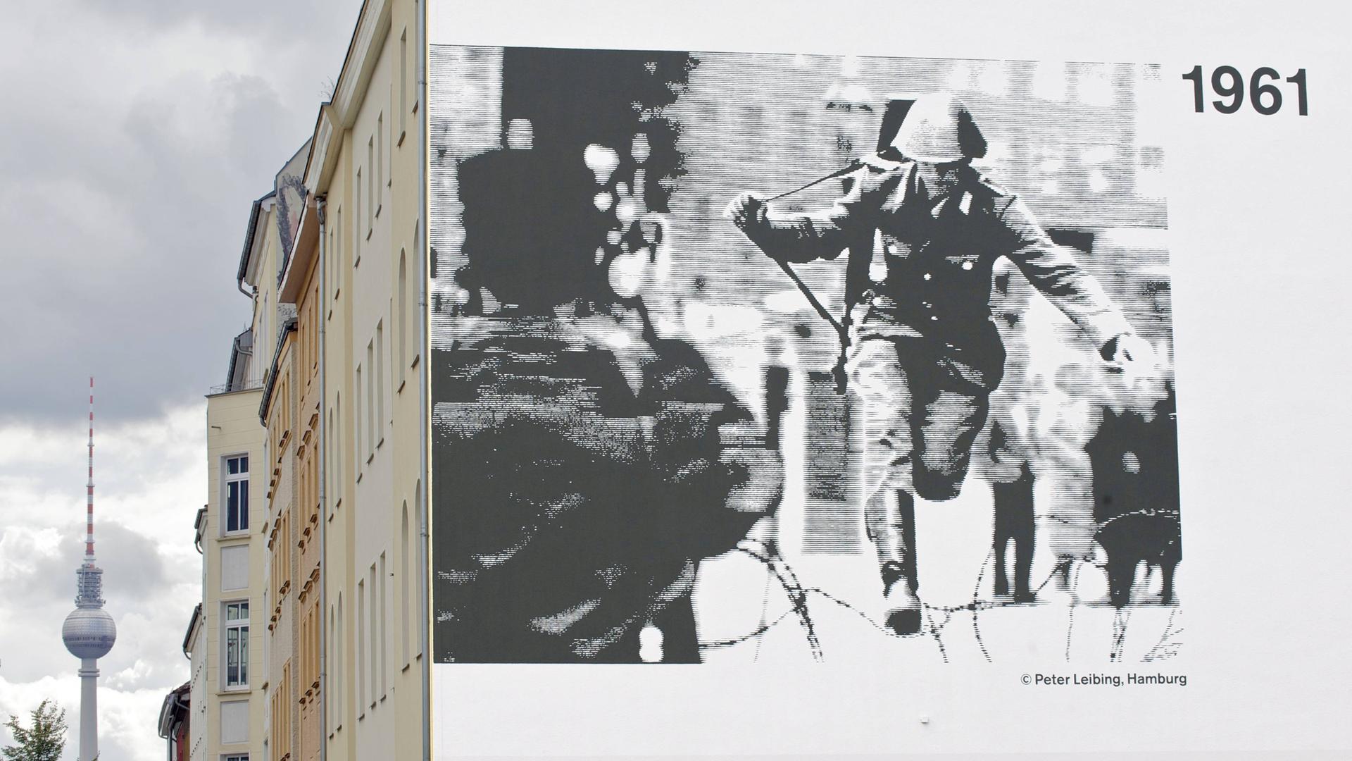 Auf einer Hauswand prangt ein Schwarz-Weiß-Foto, darauf zu sehen: Ein Polizist mit Geweht, der über einen Stacheldraht springt. Auf einer Hauswand prangt ein Schwarz-Weiß-Foto, darauf zu sehen: Ein Polizist mit Geweht, der über einen Stacheldraht springt.