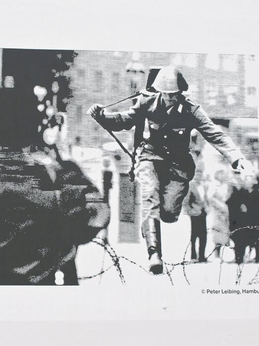 Auf einer Hauswand prangt ein Schwarz-Weiß-Foto, darauf zu sehen: Ein Polizist mit Geweht, der über einen Stacheldraht springt.