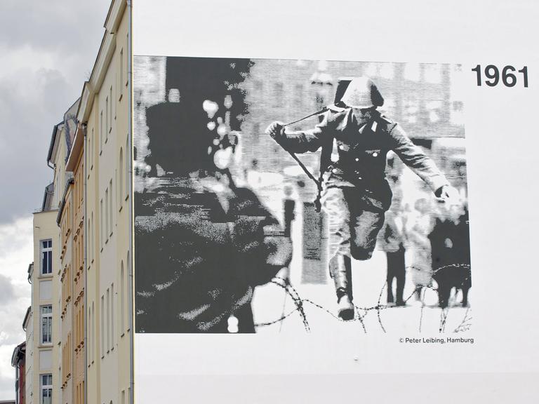 Auf einer Hauswand prangt ein Schwarz-Weiß-Foto, darauf zu sehen: Ein Polizist mit Geweht, der über einen Stacheldraht springt.