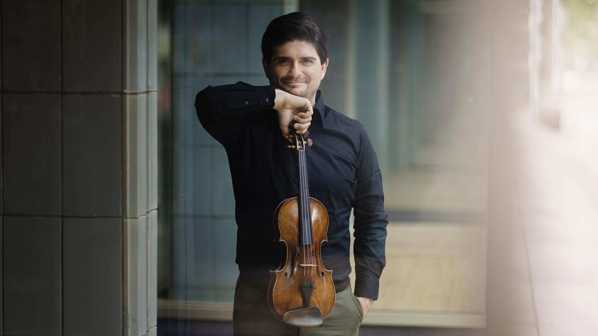 Fedor Rudin steht in einem sonnendurchflutenen Foyer mit vielen im Hintergrund immer kleiner werdenden Türen und präsentiert lächelnd seine Geige.