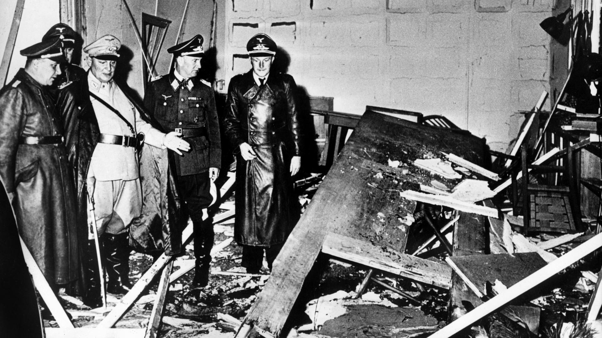 Schwarzweißfoto, das den zerstörten Raum nach dem Attentat zeigt: Reichsmarschall Hermann Göring (helle Uniform) und der Chef der "Kanzlei des Führers", Martin Bormann (l.), besichtigen den Raum der Karten-Baracke im Führerhauptquartier Rastenburg.