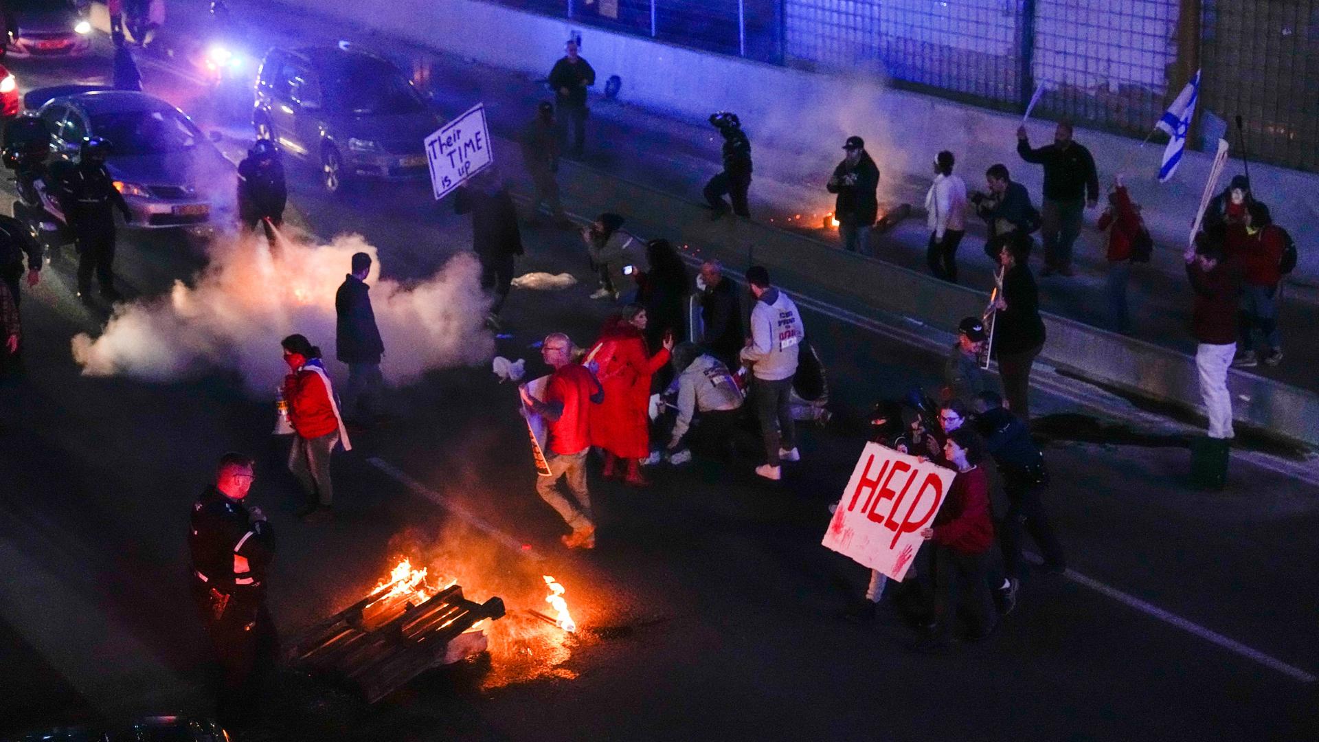 In der Dunkelheit stehen Menschen auf einer Straße. Sie halten Schilder hoch, auf denen die Freilassung der von der Hamas entführten Geiseln gefordert wird. Einige halten Fahnen. Ein Feuer brennt.