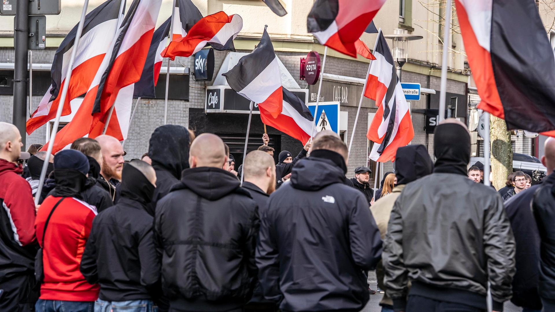 Mehrere Teilnehmer einer Demonstration der Neonazi-Gruppe "Jung und Stark" stehen mit szenetypischen Kleidungstücken nebeneinander. Im Hintergrund sind schwarz-weiß-rote Fahnen zu sehen. Mehrere Teilnehmer einer Demonstration der Neonazi-Gruppe "Jung und Stark" stehen mit szenetypischen Kleidungstücken nebeneinander. Im Hintergrund sind schwarz-weiß-rote Fahnen zu sehen.