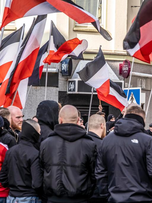 Mehrere Teilnehmer einer Demonstration der Neonazi-Gruppe "Jung und Stark" stehen mit szenetypischen Kleidungstücken nebeneinander. Im Hintergrund sind schwarz-weiß-rote Fahnen zu sehen.